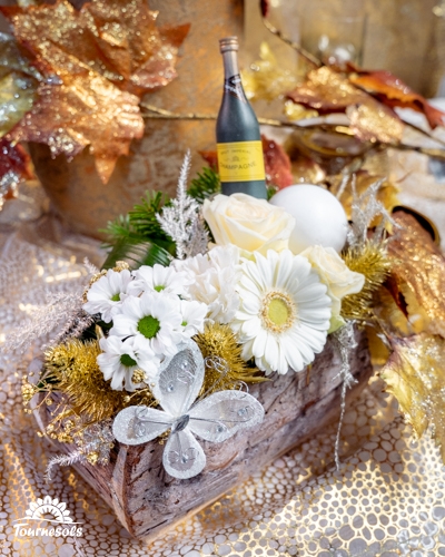 Composition florale Nouvel An blanc et or avec gerbera, roses, feuillage doré et mini bouteille festive.